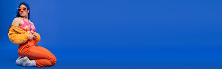 fashion statement, full length of female model with blue hair and trendy sunglasses sitting on blue background, rebel style, modern fashion, trendy accessory, youth, young beautiful woman, bannerの写真素材