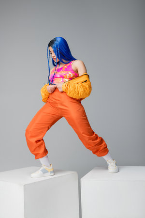 full length, fashion forward, young woman with blue hair posing in puffer jacket and orange pants on...