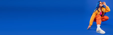generation z, full length of pretty female model with blue hair and trendy sunglasses sitting on haunches on blue background, rebel style, modern fashion, trendy accessory, bannerの写真素材