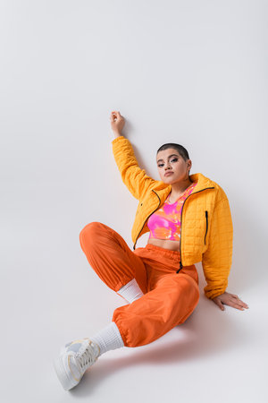 colorful clothes, outerwear, female model posing in casual attire, young woman with short hair and puffer jacket sitting on grey background, looking at camera, personal style, self expressionの写真素材