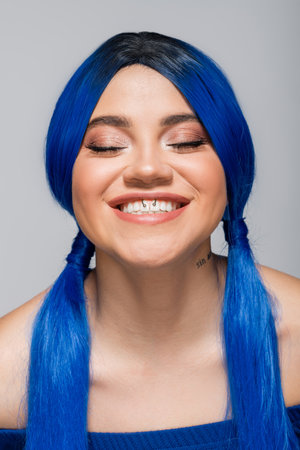 positivity and youth, tattooed woman with closed eyes and blue hair smiling on grey background,...