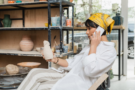 Side view of young asian artist in headscarf and workwear talking on smartphone and holding ceramic sculpture while sitting in ceramic workshop, artisan in pottery studio focusing on creationの写真素材