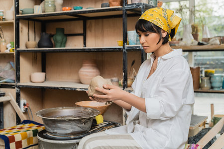 Young asian female artisan in headscarf and workwear holding clay while working near pottery wheel in ceramic workshop at background, craftsmanship in pottery makingの写真素材