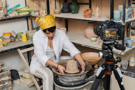 Young asian female artisan in workwear and headscarf molding clay on pottery wheel near blurred digital camera on tripod in ceramic workshop, pottery artist showcasing craftの写真素材