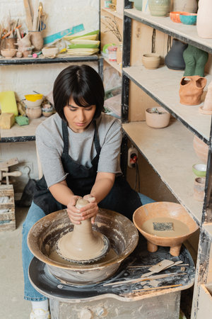 Young asian female artisan in apron molding wet clay on pottery wheel near bowl with water and tools while working in ceramic workshop, pottery tools and equipmentの写真素材