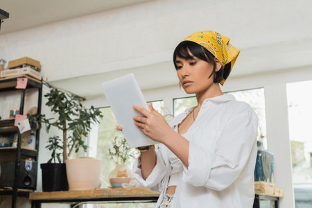 Young brunette asian female artist in headscarf and workwear using digital tablet while working in...