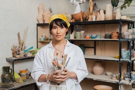 Young brunette asian female artisan in workwear and headscarf holding pottery tools and looking at camera while standing in ceramic workshop, creative process of pottery makingの写真素材