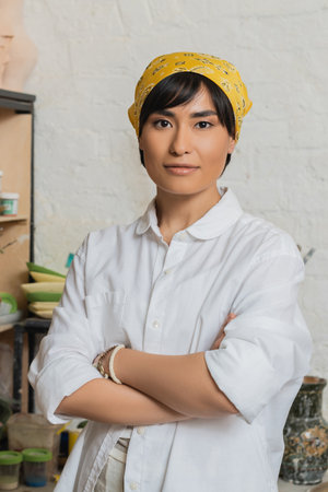 Portrait of young asian female potter in headscarf and workwear crossing arms and looking at camera while standing in ceramic workshop, pottery studio scene with skilled artisanの写真素材