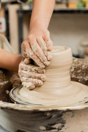 Cropped view of young female ceramicist molding wet clay on spinning pottery wheel while working in blurred ceramic workshop at background, skilled pottery making conceptの写真素材
