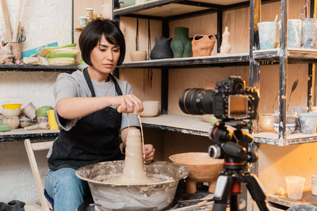 Young brunette asian female artisan in apron pouring water from sponge on wet clay and pottery wheel near digital camera on tripod in studio at background, pottery tools and equipmentの写真素材