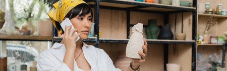 Young asian female potter in headscarf and workwear talking on smartphone and holding clay sculpture while working in ceramic workshop, artisan in pottery studio focusing on creation, bannerの写真素材