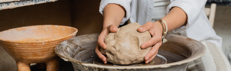 Cropped view of female artist in workwear putting clay on pottery wheel near bowl while working in...