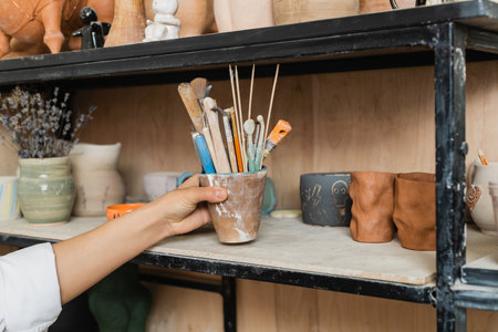 Cropped view of young craftswoman taking pottery tools from shelf with clay products and blurred lavender flowers at background in ceramic workshop, creative process of pottery makingの写真素材