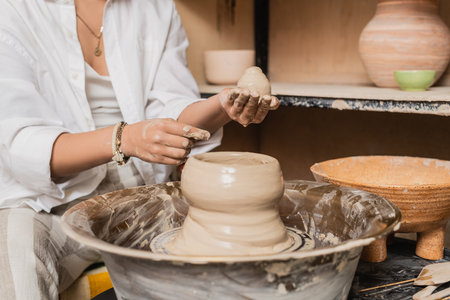 Cropped view of young female artisan in workwear holding and molding clay on pottery wheel and working near bowl in ceramic workshop, clay sculpting process conceptの写真素材
