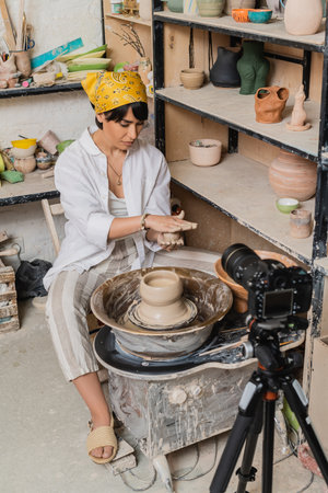 Young asian artisan in headscarf and workwear holding wet clay and working near pottery wheel and digital camera on tripod in ceramic studio, clay sculpting process conceptの写真素材