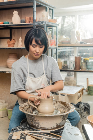 Young asian female artist in apron and workwear creating clay jug on pottery wheel near wooden tools on table in blurred ceramic workshop at background, clay shaping technique and processの写真素材