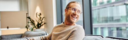 stylish and optimistic businessman in eyeglasses and casual clothes sitting in lounge of modern coworking office, smiling and looking away through window, business success concept, bannerの写真素材