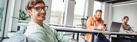 successful and optimistic team lead in stylish eyeglasses smiling at camera in modern office...