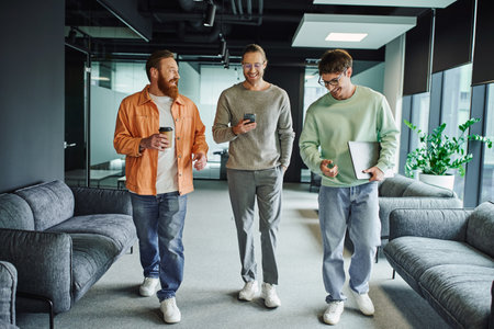 cheerful entrepreneurs in stylish casual clothes walking with mobile phone, laptop and takeaway...