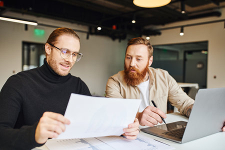 smiling entrepreneur in black turtleneck and eyeglasses looking at paper near surprised bearded business partner, charts and laptop on work desk in contemporary office, startup planning conceptの写真素材