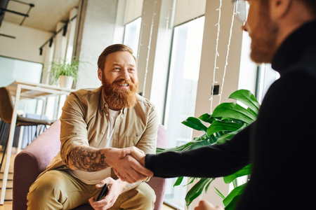 cheerful, bearded and tattooed businessman shaking hands with ambitious entrepreneur in black turtleneck while sitting in contemporary office, successful partnership conceptの写真素材