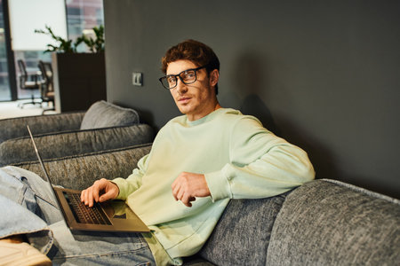 ambitious entrepreneur in stylish eyeglasses and casual clothes sitting on comfortable couch with laptop and looking at camera in contemporary office environment, confidence and success conceptの写真素材