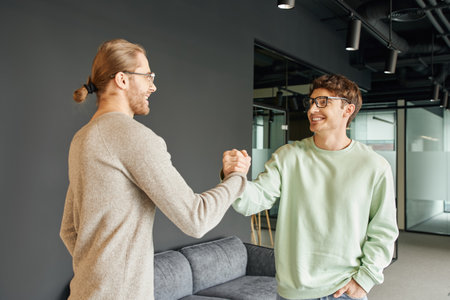 cheerful and stylish entrepreneurs in eyeglasses and casual clothes shaking hands and confirming agreement in lounge of modern coworking office, successful collaboration conceptの写真素材