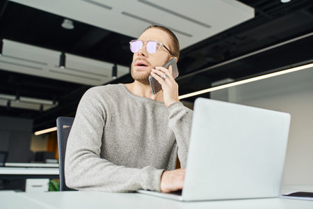 thoughtful businessman in eyeglasses looking away and talking on mobile phone while sitting near laptop on work desk, startup project planning in contemporary office spaceの写真素材