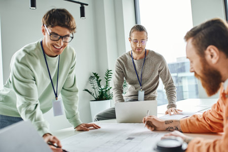 smiling architect pointing at blueprint near colleagues and laptop while working in coworking space of architectural studio, business collaboration and implementation of innovative ideasの写真素材