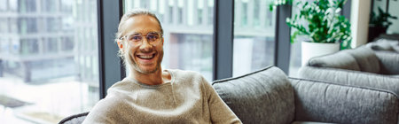 professional headshot of smiling and successful businessman in casual clothes and stylish eyeglasses sitting on comfortable couch in lounge of modern coworking office and looking at camera, bannerの写真素材