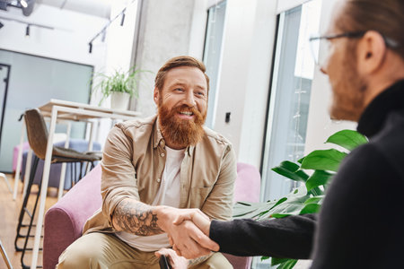overjoyed, tattooed and bearded entrepreneur in casual clothes confirming agreement and shaking hands with businessman on blurred foreground, successful partnership conceptの写真素材