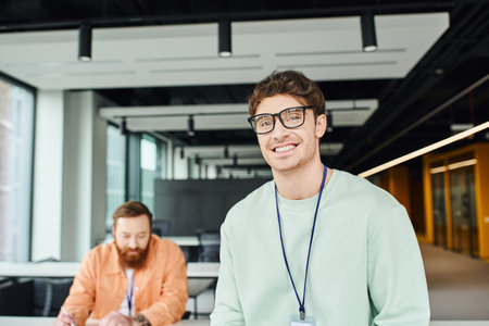cheerful and optimistic architectural designer in eyeglasses smiling at camera near bearded colleague working on business startup on blurred background in modern office environmentの写真素材