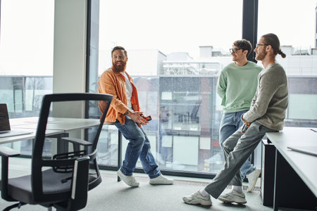 positive entrepreneurs in stylish casual clothes smiling while standing near work desks and large...