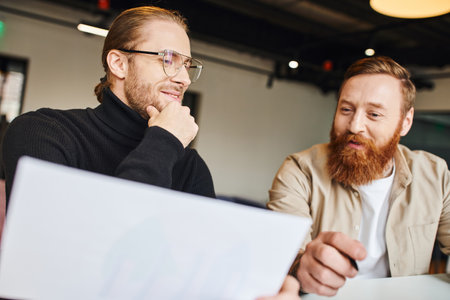 pleased businessman in black turtleneck and eyeglasses touching chin while looking at document near happy bearded colleague while working on new startup in coworking officeの写真素材