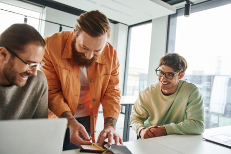 bearded and tattooed designer showing color samples to cheerful and stylish entrepreneurs in eyeglasses sitting near laptops in contemporary coworking environment, teamwork and business creativityの写真素材