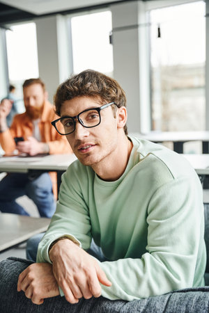 positive and ambitious entrepreneur in eyeglasses and casual clothes looking at camera while his...