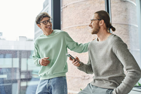 joyful entrepreneur in eyeglasses and casual clothes touching shoulder of business partner sitting with smartphone and hand in pocket near large office window, concept of successful collaborationの写真素材