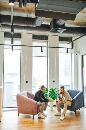 full length of bearded man showing smartphone to colleague in black turtleneck while sitting in...