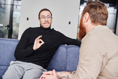 businessman in eyeglasses and black turtleneck showing okay sign while planning startup project with bearded tattooed colleague in modern office, business collaboration conceptの写真素材