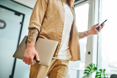 partial view of tattooed entrepreneur in stylish casual clothes standing with laptop and messaging on mobile phone in modern office environment, successful business conceptの写真素材