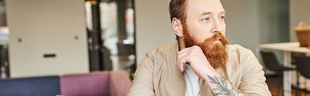 portrait of thoughtful, bearded and tattooed entrepreneur looking away in modern office environment,...