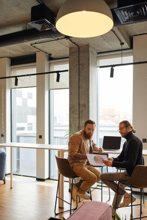 businessman in eyeglasses and black turtleneck looking at charts with bearded and tattooed colleague...