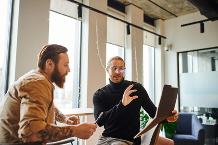 inspired businessman in eyeglasses and black turtleneck showing folder with startup planning to bearded colleague in contemporary coworking office, productivity and collaboration conceptの写真素材