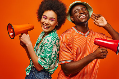 Positive young african american best friends in summer clothes looking at camera while holding loudspeakers and standing isolated on red, friends in fashionable outfits, friendshipの写真素材