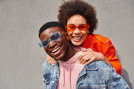 Portrait of cheerful young african american woman with natural hair embracing best friend in stylish sunglasses and denim jacket while looking at camera near building, trendy friends in urban settingsの写真素材