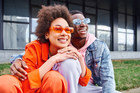 Pleased young african american woman in sunglasses and bright outfit looking away while spending time with best friend and sitting on blurred urban street, friends with trendy aestheticの写真素材