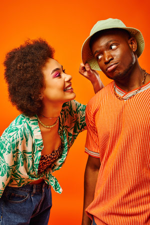 Cheerful young african american woman with bold makeup touching panama hat of trendy best friend while spending time isolated on orange, friends showcasing individual style, friendshipの写真素材