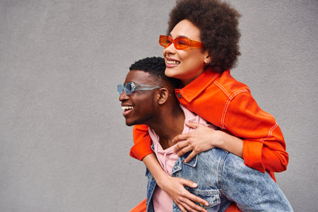 Young and stylish african american woman in sunglasses piggybacking on best friend in denim jacket and having fun near building on urban street, trendy friends in urban settingsの写真素材