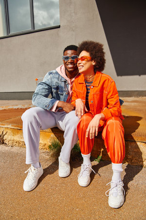 Smiling young african american man in sunglasses hugging trendy best friend in bright outfit while sitting on rusty stairs near building on urban street, stylish friends in cityの写真素材