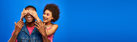 Smiling young african american woman with bright makeup covering eyes of trendy best friend in denim...